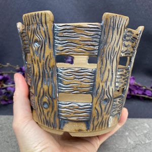 May include: A handcrafted ceramic planter with a rustic, wood-like design. The planter features a brown and blue color scheme, with carved details mimicking wooden planks and knots. The open design allows for air circulation and drainage, perfect for small plants.