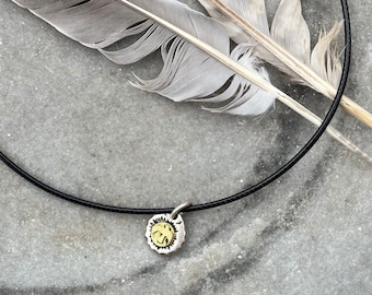 May include: A black leather cord necklace with a silver and gold pendant. The pendant is round and has a textured surface. The necklace is lying on a gray surface with a feather.