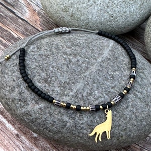 Puede incluir: Un brazalete de cuentas negras y doradas con un dije de lobo. El brazalete está hecho con pequeñas cuentas negras, cuentas doradas y cuentas rayadas en blanco y negro. El dije de lobo es dorado y cuelga del brazalete.