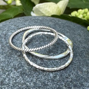 Könnte beinhalten: Drei silberne Ringe. Ein Ring hat ein gedrehtes Seildesign, und die anderen beiden haben eine gehämmerten Textur. Die Ringe werden auf einer grauen Steinoberfläche mit weißen Blumen und grünen Blättern im Hintergrund präsentiert.