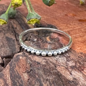 Op de afbeelding: Een zilveren ring met een kralenontwerp. De ring is dun en heeft kleine, gelijkmatig verdeelde kralen langs de band. De ring ligt op een getextureerd, bruin houten oppervlak, met groene en gele plantenelementen op de achtergrond.