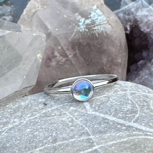 Pode incluir: Um anel de prata com uma pedra preciosa redonda e iridescente. O anel está sobre uma pedra cinza, com outras pedras no fundo. A pedra preciosa reflete tons de azul, verde e roxo.
