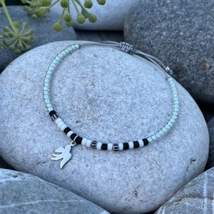 Puede incluir: Un brazalete delicado con un dije de paloma de plata. El brazalete está hecho con cuentas de color azul claro y negro y un cordón gris.