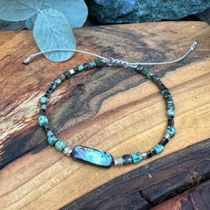 Può includere: Bracciale regolabile con perline con pietre color turchese, perline nere e un elemento centrale rettangolare in conchiglia di abalone. Il bracciale è infilato su un cordoncino grigio e poggia su una superficie di legno.