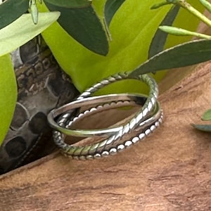 Könnte beinhalten: Drei silberne Ringe mit unterschiedlichen Texturen: ein gedrehter Seilring, ein Ring mit Perlenmuster und ein glatter Ring. Die Ringe werden auf einer Holzoberfläche mit grünem Laub im Hintergrund präsentiert.