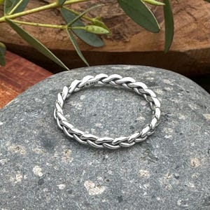 Könnte beinhalten: Ein silberner Ring mit geflochtenem Design, der auf einem grauen Stein liegt. Das geflochtene Muster des Rings ist sichtbar, und das Metall hat eine helle, reflektierende Qualität. Der Ring ist ein Schmuckstück.