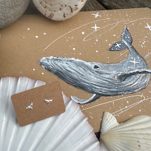 May include: A pair of silver whale tail earrings on a brown card with a painted image of a whale in the background.
