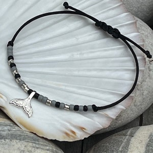 Könnte beinhalten: Ein Armband aus schwarzem Band mit grauen und silbernen Perlen und einem silbernen Wal-Schwanz-Anhänger.