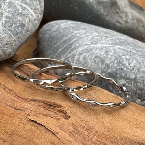 Può includere: Tre anelli d'argento con diverse trame. Uno presenta un motivo geometrico, un altro un design ondulato e il terzo è una fascia semplice. Gli anelli sono presentati su un pezzo di legno, con pietre grigie sullo sfondo.