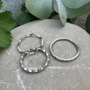 Könnte beinhalten: Ein Set aus drei silberfarbenen Ringen. Ein Ring hat eine glatte, gewellte Ringschiene. Ein zweiter Ring weist einen texturierten, gewellten Rand auf. Der dritte Ring ist ein dünner Bandring mit einem diagonalen, sich wiederholenden Muster.