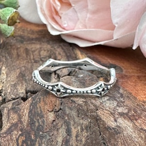 May include: A silver ring with a unique, wavy design. The ring features small star-shaped accents and tiny beaded details along the edges. The ring is displayed on a textured, wooden surface.