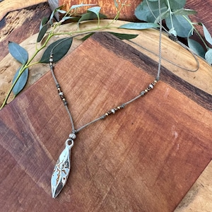Puede incluir: Un collar con colgante plateado con un diseño en forma de hoja. El colgante cuelga de un cordón gris con pequeñas cuentas doradas y plateadas. El collar se exhibe sobre una superficie de madera con follaje verde.