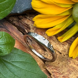 May include: A silver-toned metal ring featuring a series of engraved sunburst motifs along the outer band. The ring has a polished, reflective surface and a rounded profile, showcasing detailed line work that radiates from the center of each sun.