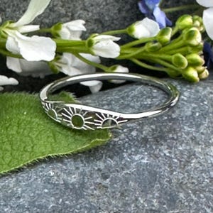 May include: A silver ring with a sunburst design. The ring has three sun motifs, each with radiating lines. The ring is shown on a green leaf and a grey stone surface, with white and blue flowers in the background.