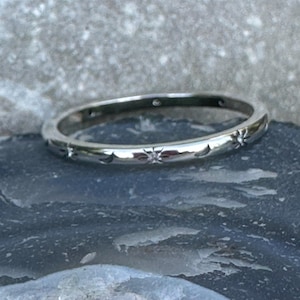 Op de afbeelding: Een zilveren ring met een herhalend patroon van sterren en halve manen. De ring heeft kleine, sprankelende accenten en een gepolijste afwerking. De ring is een delicaat sieraad.