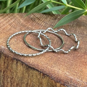 May include: A set of three silver-toned rings. One ring features a textured, faceted surface, while the second ring has a smooth, plain band. The third ring displays a distinct wavy, undulating design. All three rings are thin and circular.