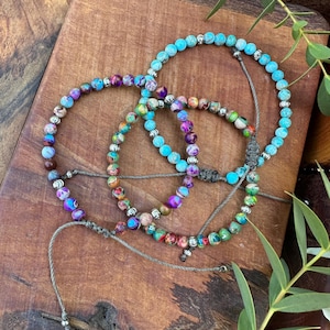 Könnte beinhalten: Drei verstellbare Armbänder mit runden, mehrfarbigen Jaspisperlen in Blau-, Lila-, Grün- und Brauntönen, durchsetzt mit kleinen silberfarbenen Metallzwischenperlen. Jedes Armband verfügt über eine graue Kordel mit einem Schiebeknotenverschluss.