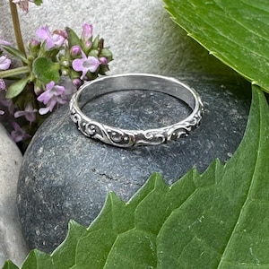 Könnte beinhalten: Ein silberner Ring mit einem kunstvollen, wirbelnden Muster. Der Ring liegt auf einem grauen Stein, mit grünen Blättern und kleinen lila Blumen im Hintergrund. Das Design des Rings zeigt ein sich wiederholendes Muster aus Kurven und Schnörkeln.