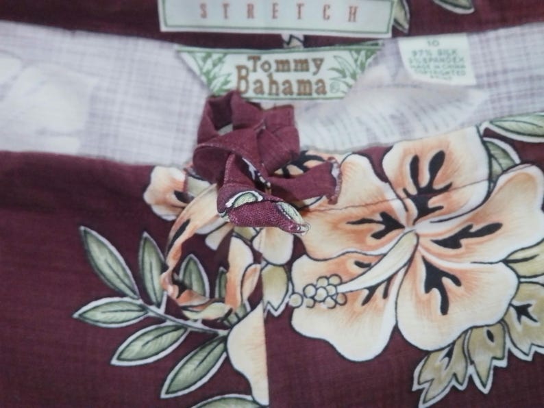 May include: A maroon and white patterned fabric with a floral design. The fabric has a brown drawstring with a bow. The fabric has a label that says "Tommy Bahama" and "Stretch".