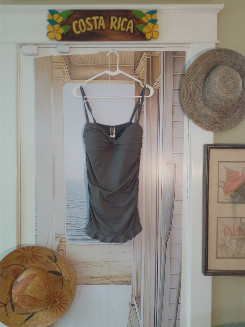 May include: A grey tank top with a ruffled hem hangs on a white hanger against a backdrop of a beach scene. The tank top has a strapless design and a gathered bodice.