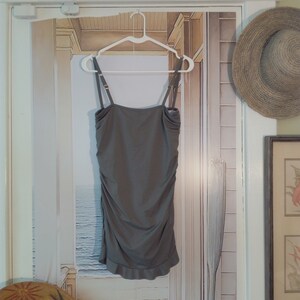 May include: A grey sleeveless dress with a ruffled hem, hanging on a white hanger against a backdrop of a painted beach scene. The dress has a simple, relaxed fit and a slightly gathered bodice.