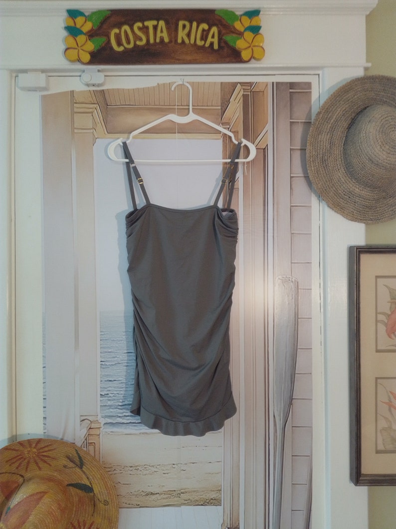 May include: A grey sleeveless dress with a ruffled hem, hanging on a white hanger against a backdrop of a painted beach scene. The dress has a simple, relaxed fit and a slightly gathered bodice.