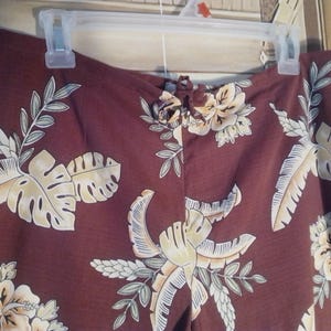 May include: A pair of brown pants with a tropical floral print featuring large leaves and flowers in shades of yellow, white, and green.