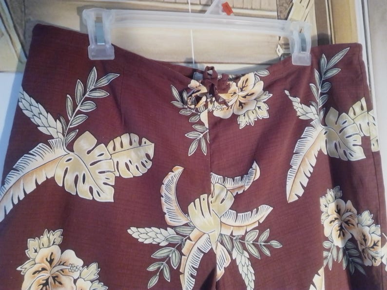May include: A pair of brown pants with a tropical floral print featuring large leaves and flowers in shades of yellow, white, and green.