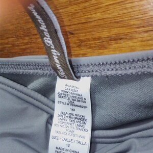 May include: A grey swimsuit tag with the brand name Tommy Bahama, style number TSW64623P, size 12, and fabric composition details. The tag also includes the text "Made in China, Fait aux Chine, Hecho en China".