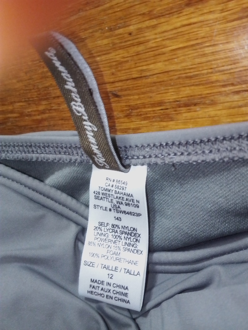 May include: A grey swimsuit tag with the brand name Tommy Bahama, style number TSW64623P, size 12, and fabric composition details. The tag also includes the text "Made in China, Fait aux Chine, Hecho en China".