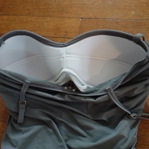 May include: A grey strapless swimsuit with white padding in the bust area. The swimsuit has adjustable straps and a back closure.