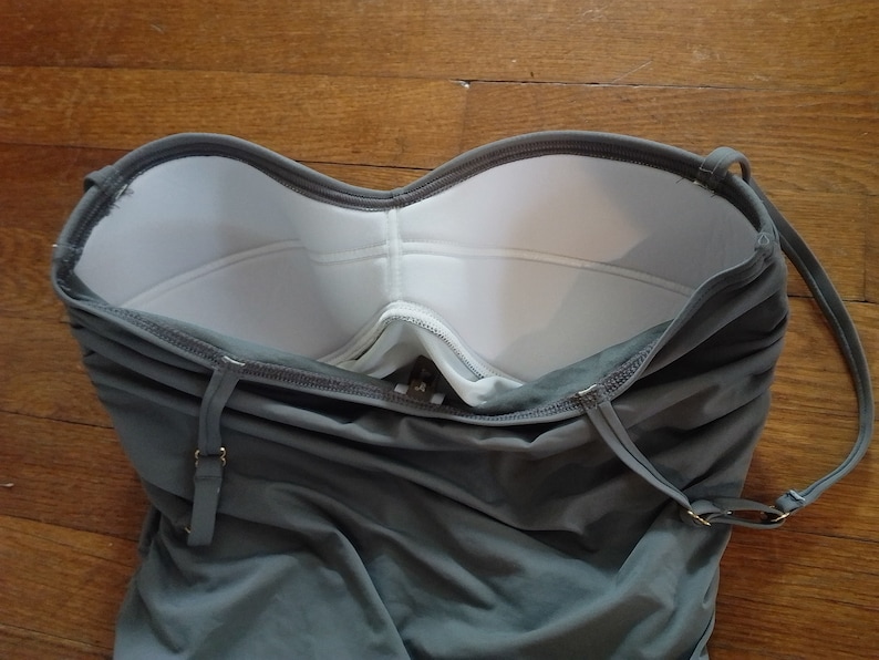 May include: A grey strapless swimsuit with white padding in the bust area. The swimsuit has adjustable straps and a back closure.