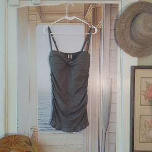 May include: A grey strapless swimsuit top with a ruffled hem. The top is hanging on a white hanger against a backdrop of a beach scene.