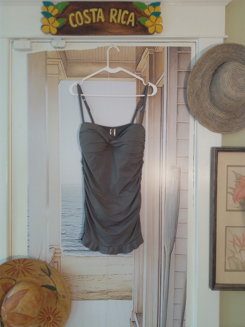 May include: A grey strapless swimsuit top with a ruffled hem. The top is hanging on a white hanger against a backdrop of a beach scene.