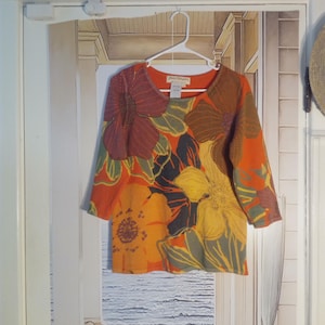 May include: An orange, long-sleeved shirt with a floral pattern in shades of yellow, brown, and green. The shirt is hanging on a white hanger. A wooden sign that says "COSTA RICA" is above the shirt.
