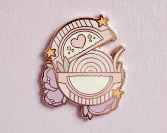 Puede incluir: Un pin de esmalte rosa y dorado con una ilustración de dibujos animados de un tazón de fideos con un corazón en la tapa, rodeado de flores y estrellas.