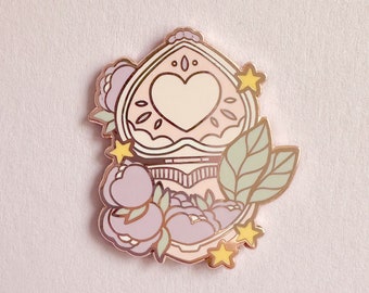 Puede incluir: Un pin de esmalte dorado con un diseño de un compacto en forma de corazón con detalles florales y estrellas. El compacto está abierto, revelando un corazón rosa en su interior. El pin tiene una paleta de colores pastel.