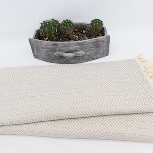 Pode incluir: Uma manta de arremesso bege com um padrão de espinha de peixe e bordas com franjas. A manta está dobrada ao meio e está sobre uma superfície branca.