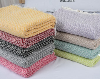 Pode incluir: Uma pilha de oito mantas de algodão tecidas em várias cores, incluindo mostarda, bordô, preto, cinza, fúcsia, lilás, bege, menta e verde pistache. As mantas apresentam um padrão chevron e estão dispostas em forma escalonada. Est. 2015 está impresso no fundo.