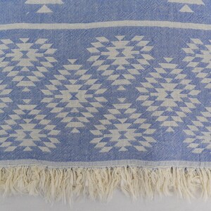 Peut inclure: Une couverture en coton bleu et blanc avec une bordure en franges. Le motif est un design g&eacute;om&eacute;trique de triangles et de carr&eacute;s.