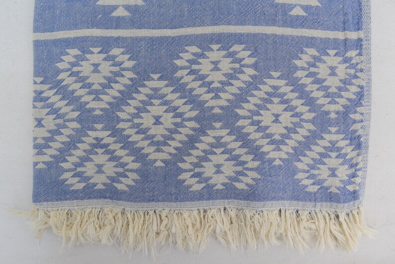 Peut inclure: Une couverture en coton bleu et blanc avec une bordure en franges. Le motif est un design g&eacute;om&eacute;trique de triangles et de carr&eacute;s.