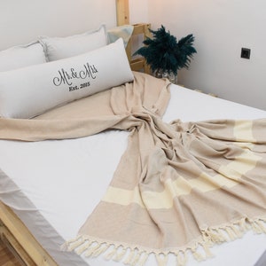 May include: A white bed with a beige and white striped throw blanket with tassels. The bed has a white pillow with the text "Mr. & Mrs. Est. 2005" on it.