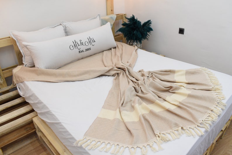 May include: A white bed with a beige and white striped throw blanket with tassels. The bed has a white pillow with the text "Mr. & Mrs. Est. 2005" on it.