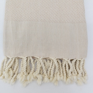May include: A cream-coloured Turkish towel with a woven pattern and fringe. The towel is made of cotton and has a soft, absorbent texture.