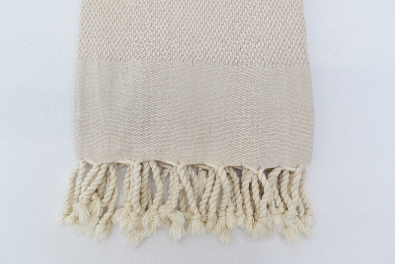 May include: A cream-coloured Turkish towel with a woven pattern and fringe. The towel is made of cotton and has a soft, absorbent texture.