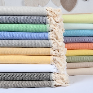 Peut inclure: Une pile de 14 serviettes turques de différentes couleurs, notamment gris clair, bleu marine, vert, bleu clair, gris foncé, moutarde, jaune foncé, blanc, noir, marron, vert pâle, turquoise, violet, orange, jaune et vert kaki. Les serviettes sont pliées et empilées les unes sur les autres, avec les franges de chaque serviette visibles.