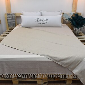 May include: A beige throw blanket with fringe on a bed with white pillows. The pillows have the text "Mr. & Mrs. Est. 2015" embroidered on them.