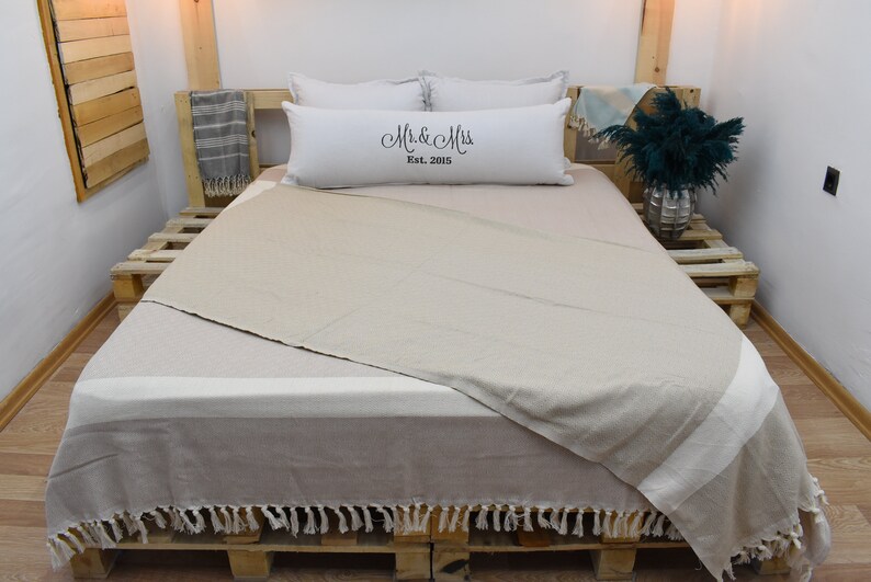 May include: A beige throw blanket with fringe on a bed with white pillows. The pillows have the text "Mr. & Mrs. Est. 2015" embroidered on them.