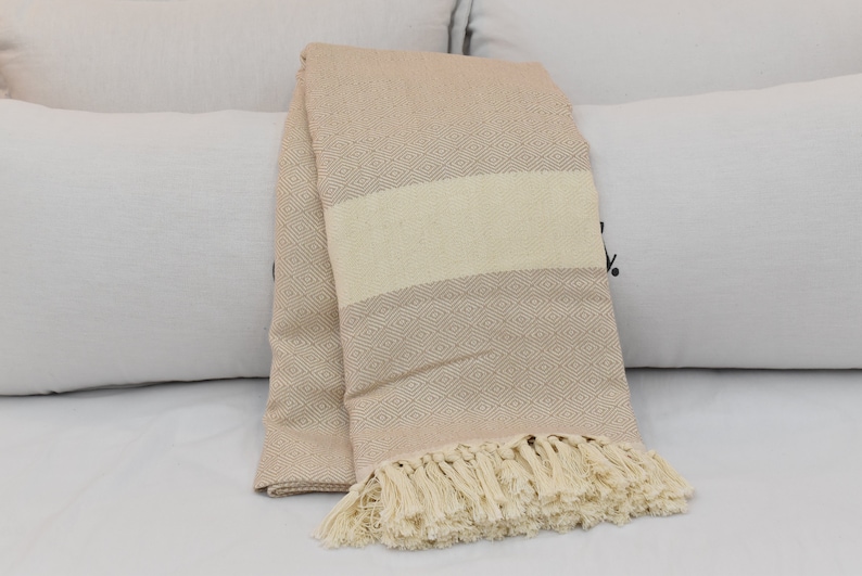 May include: A beige throw blanket with a diamond pattern and a white stripe. The blanket has a fringe border.