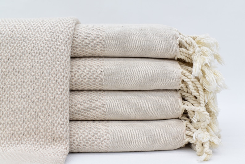 May include: A stack of five cream-coloured Turkish towels with fringed edges. The towels are made of cotton and have a woven texture.
